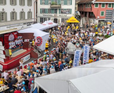 Dorfplatz voller Menschen, sammeln sich um eine Bühne, auf der die Fahrer sich präsentieren